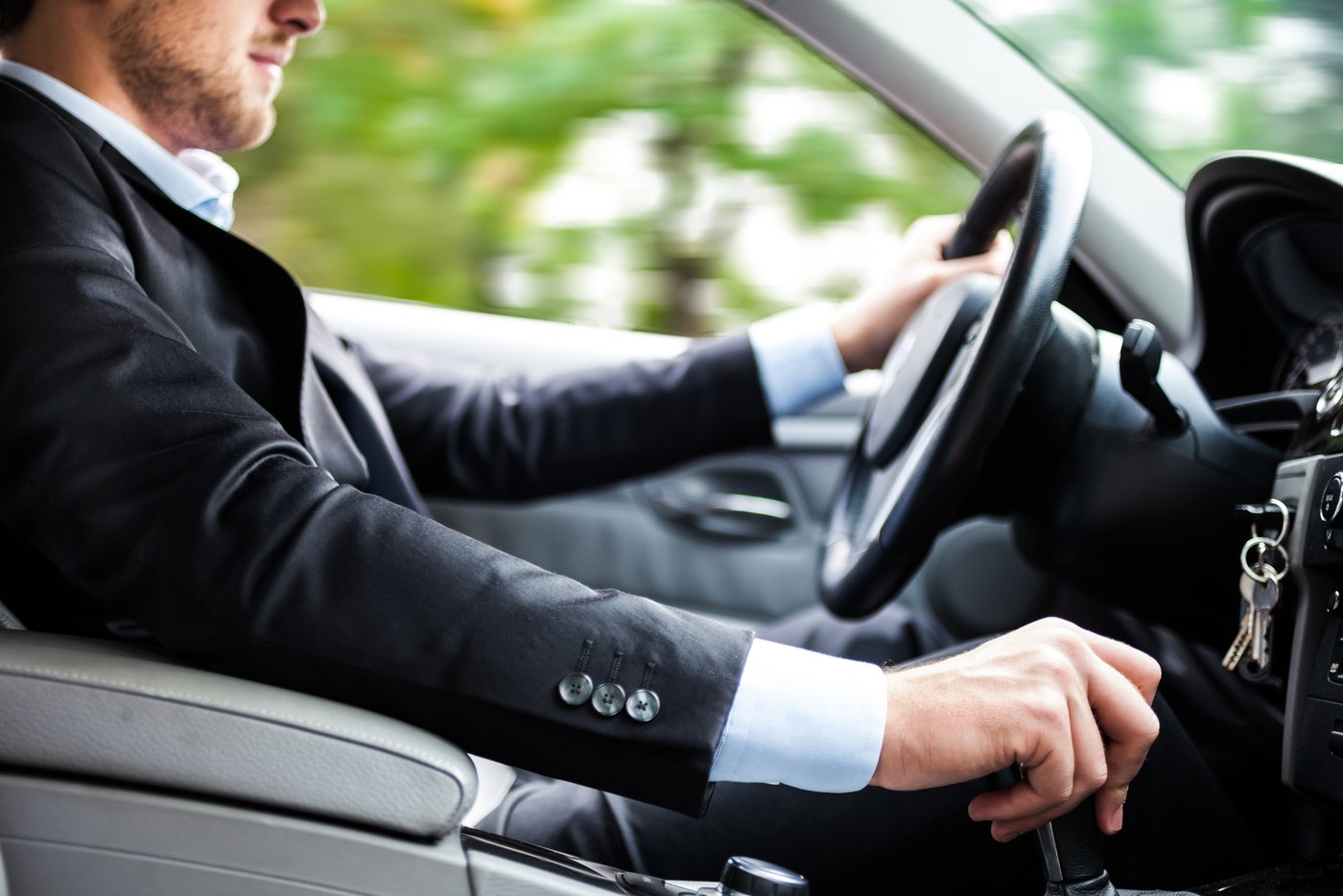 Uomo in abito scuro e camicia azzurra al volante di un'auto, mano sul cambio manuale e chiavi inserite nel cruscotto